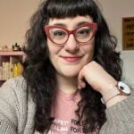 This picture is of Rachel Harlich, LCSW. The image is of a white non-binary femme person with dark brown curly hair and bangs wearing a light pink t-shirt that says "healing for everyone" and other text that is too small to read. She is also wearing a taupe sweater and is propping her head up with her hand. She is wearing a watch. Behind her are bookshelves and posters.